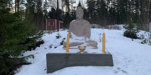 Eine graue Buddha-Statue steht auf einem Sockel vor zwei gelben Kerzen und einem Gefäß in einem schneebedeckten Wald. Im Hintergrund steht eine rote Holzhütte.