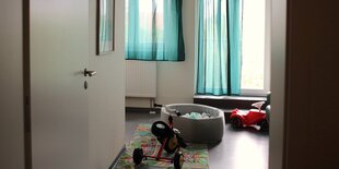 Auf dem Bild ist eine Wohnung zu sehen. Im Flur steht ein Dreirad und ein kleines Bällebad. Vor den Fenstern hängen durchscheindende türkise Gardinen.