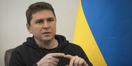 Michailo Podoljak sitzt vor einer ukrainischen Flagge