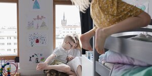 Ein Kind hängt von einem Stockbett. Im Hintergrund ein Mädchen und ein Junge die gemeinsam lesen