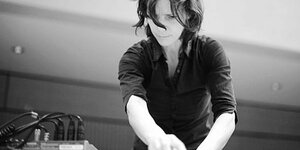 Schwarz-Weiß-Aufnahme der Echtzeitmusikerin Andrea Neumann. Sie steht an einem Mischpult und dreht an Knöpfen. Ihre Haaren fallen über ihre Stirn und sie trägt ein dunkles Hemd mit hochgekrempelten Ärmeln.