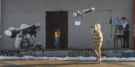 Eine Frau geht an künstlerischen Arbeiten von TvBoy vorbei die eine Granate zeigen, die ein Soldat abschiesst