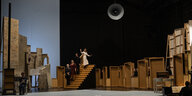 Auf einer Bühne sitzt links ein Mann an einem Tisch und hat eine Trompete in der Hand. Auf einer Treppe stehen vier weitere Schauspielerinnen und Schauspieler. Von der Decke hängt ein großes Nebelhorn