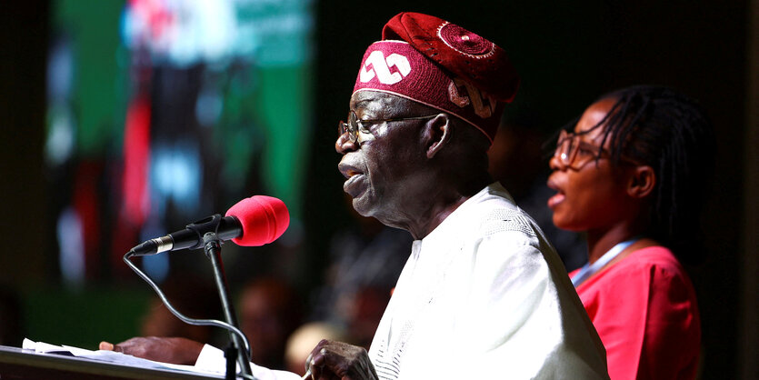 Bola Tinubu spricht in ein Mikrofon, er trägt eine rote Kopfbedeckung