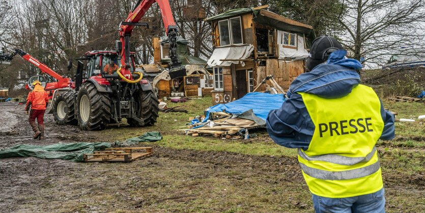 Ein Journalist mit gelber Warnweste, auf der "Presse" steht, beobachtet einen Bagger, der in Lützerath ein Haus einreißt