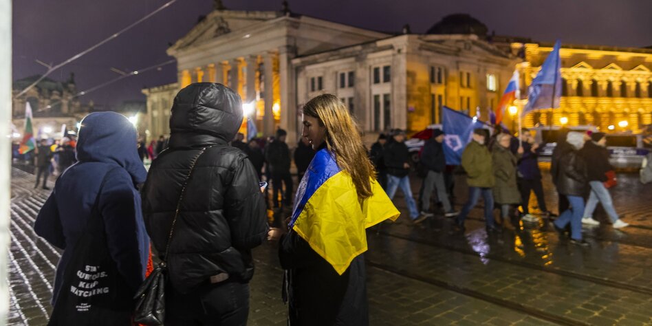 Drei Personen mit Ukrainefahne stehen abseits eines rechten Demonstrationszuges, der mit Russlandfahnen an der Semperoper vorbeimarschiert