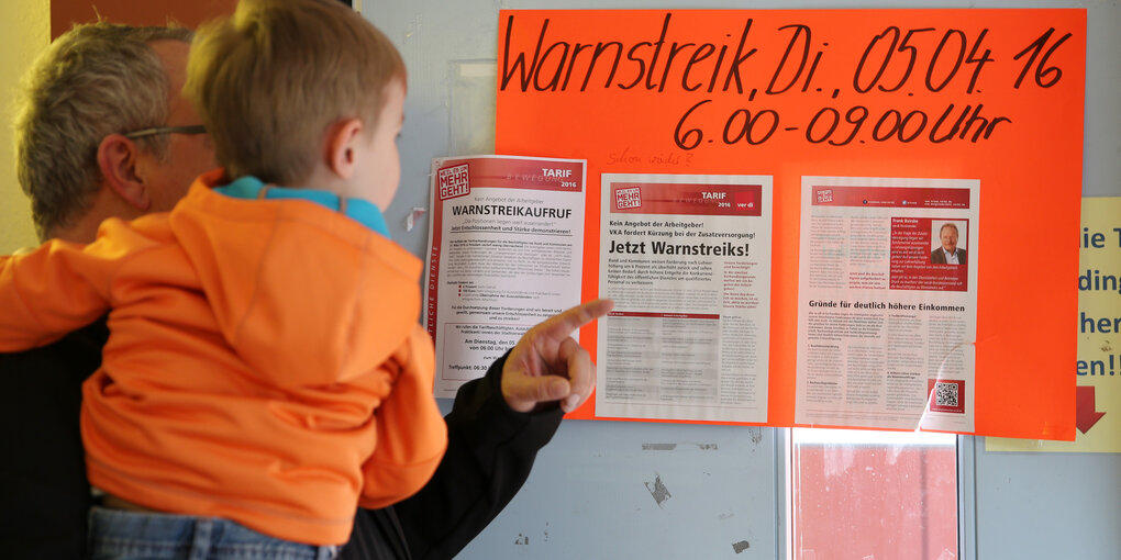 Ein Vater mit seinem kleinen Sohn auf dem Arm steht vor einem Plakat auf dem Warnstreiks in der Kita angekündigt werden.