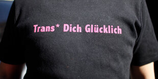 Ein Mensch trägt ein schwarzes T-Shirt mit der rosa Aufschrift "Trans* Dich Glücklich"