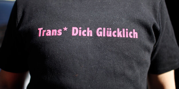 Ein Mensch trägt ein schwarzes T-Shirt mit der rosa Aufschrift "Trans* Dich Glücklich"
