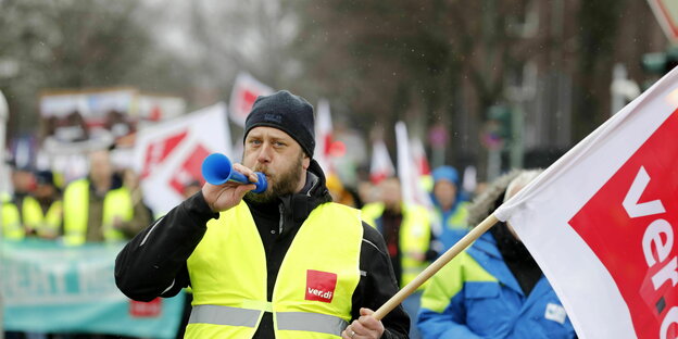 Ein Mann in einer Weste der Gewerkschaft Verdi und einer Gewerkschaftsfahne steht vor zahlreichenden Streikenden und pustet in eine Tröte