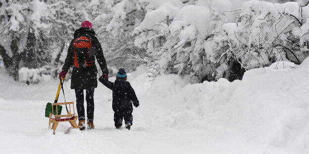 Eine Frau mit Schlitten und ein Kind gehen im Schnee und sind von hinten zu sehen