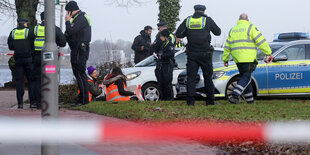 Menschen hocken vor Autos, Polizist:innen stehen daneben, im Hintergrund die Alster