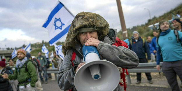 Ein israelischer Reservist, der an einer Blockade teilnimmt spricht in ein Megaphon, hinter ihm Protestierende mit israelischen Flaggen