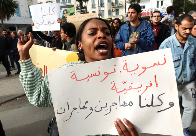 eine junge Frau auf einer Protestaktion trägt ein Schild auf dem in arabischer Sprache steht: "FeministInnen, Tunesier, Afrikaner. Wir sind alle männliche und weibliche Immigranten!