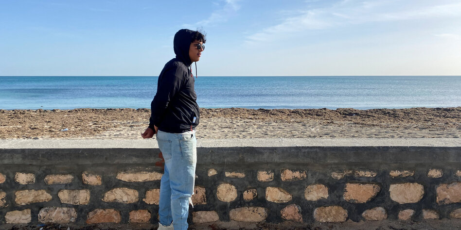 Ein junger Mann mit Hoodie steht am Strand und schaut übers Meer