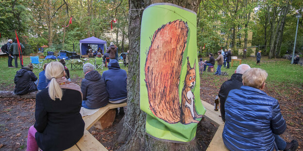 Meschen sitzen auf einer Baum rund um einen Baum, auf einem Plakat ein großes Eichhörnchen