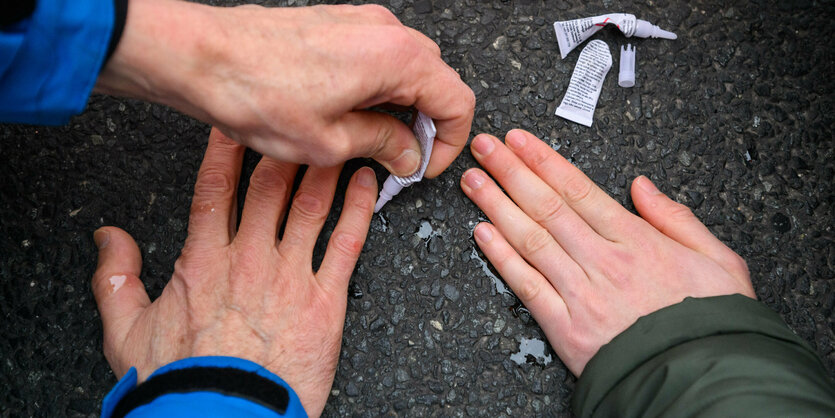 Zwei Hände kleben auf Asphalt, eine weitere Hand tropft Kleber nach.