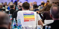 EIn Mann trägt auf seinem T-Shirt-Rücken den Spruch "Lovers Unite" mit Regenbogenstreifen