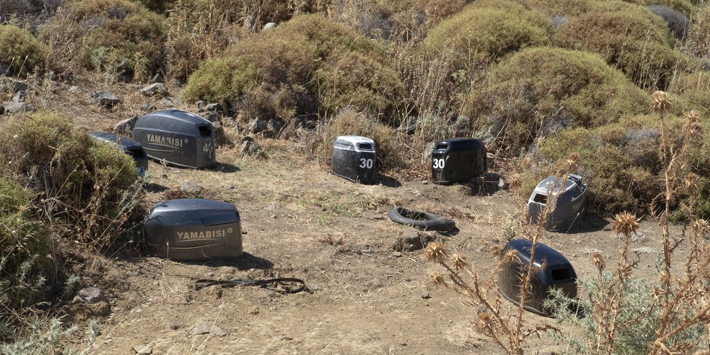 Sieben Handmotoren, wie sie bei Motorbooten eingesetzt werden, liegen auf einem sandigen Boden, der von Büschen und trockenen Gräsern umgeben ist. Einige der Motoren tragen die Aufschrift "Yamabisi"