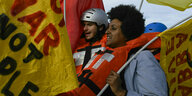 Ein Mann und eine Frau tragen Rettungswesten, sie halten Fahnen in den Händen und demonstrieren