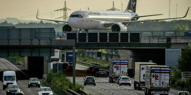 Eine Lufthansamaschine fährt über eine Brücke, die eine Autobahn kreuzt auf der Lastwagen und PKW unterwegs sind