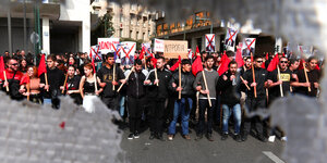 StudentInnen protestieren mit roten Fahnen und Schildern auf denen das Portrait des ehemaligen Verkehrsministers Kostas Karamanlis zu sehen ist. Sein Gesicht ist mit einem roten kreuz durchgestrichen