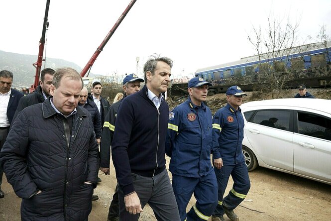 Kyriakos Mitsotakis und Kostas Karamanlis lassen sich mit ernster Miene von Feuerwehrleuten den Unglücksort zeigen