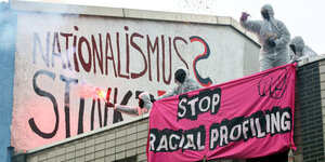 Auf einer Hauswand im Hintergrund steht "Nationalismus stinkt". Im Vordergrund stehen Aktivist*innen in weißen Staubanzügen auf einem Hausdach mit dem Banner "Stop Racial Profiling"