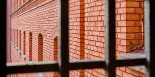 Blick aus einem vergitterten Fenster auf die Backsteinwand eines Gefängnisses