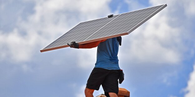 Ein Arbeiter traegt ein Solarpanel auf ein Dach eines Wohnhauses
