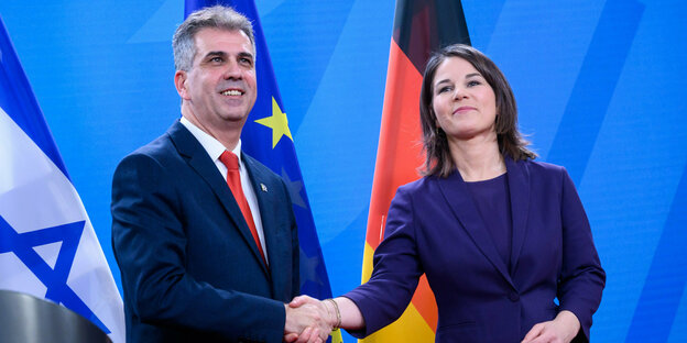 Annalena Baerbock und Eli Cohen reichen sich die Hand, im Hintergrund die Flaggen Israels, Deutschlands und der EU