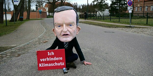Eine protestierende Person bekleidet mit einem überdimensioniertem Kopf von Volker Wissing sitzt aufeiner Dorfstrasse im Stil der letzten Generation und hat ein Schild aufgestellt: Ich verhindere Klimaschutz
