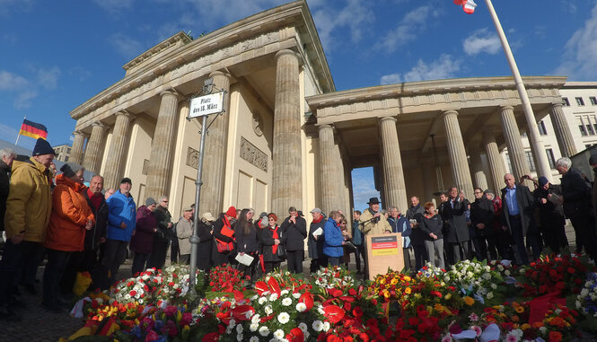 Zahlreiche Menschen gedenken auf dem Platz des 18. März am Brandenburger Tor den Opfern der März-Revolution 1848. Im Vordergrund bunte Blumensträuße.