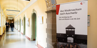 Auf einem langen Flur laufen zwei Männer der Kamera entgegen, auf einem im Vordergrund Plakat steht: Aus Niedersachsen nach Auschwitz. Die Verfrolgung der Sinti und Roma in der NS-Zeit