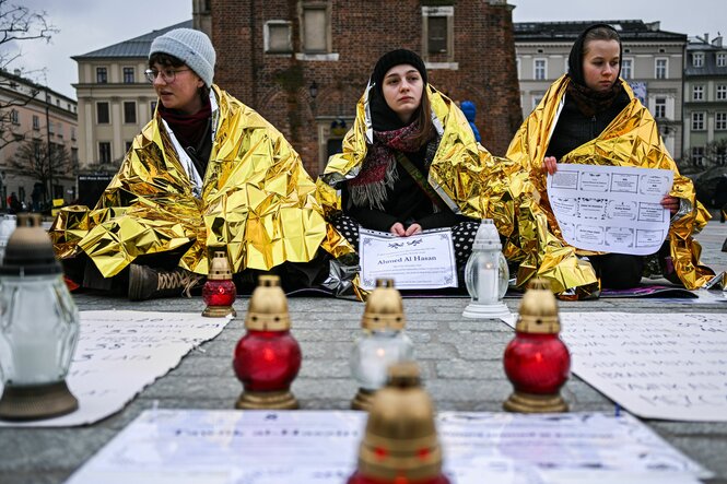 Drei junge Frauen sitzen mit goldenen Aludecken auf der Straße in Krakau mit Todesanzeigen von Geflüchteten, die in den Wäldern an der Grenze gestorben sind