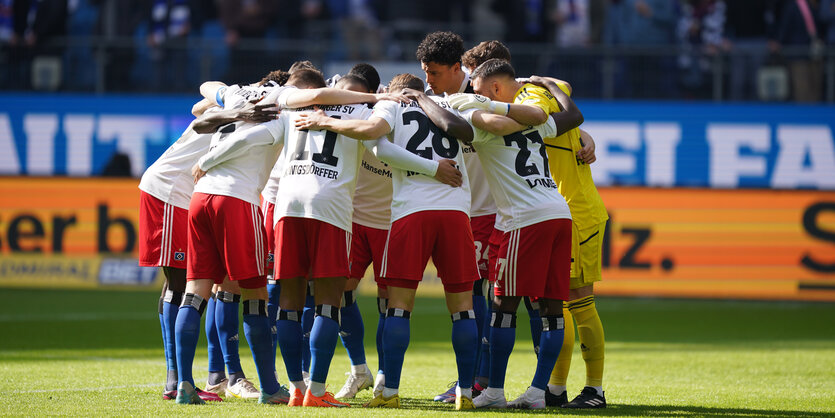 Fußballspieler in weißen Trikots und roten Hosen bilden Schulter an Schulter einen Kreis