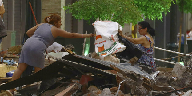 Two women hand each other a bag, with rubble between them