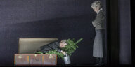 Eine Mann liegt mit einer Gießkanne und einem Tannenbaum in einem Koffer, eine Frau schaut auf ihn hinunter