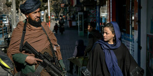 Ein bewaffneter Taliban steht Wache, während eine Frau mit Tuch über dem Haar, an ihm vorbei läuft