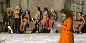 Männer und Frauen stehen in Badekleidung an einer Mauer und sonnen sich, auf dem Boden liegt Schnee, eine Frau im orangen Mantel geht vorbei