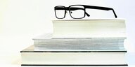 Eine Brille mit schwarzem Gestell liegt auf einem Stapel Bücher