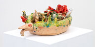 Auf einem weißen Sockel steht die bunte Keramikskulptur "Boat" von Ernie Wang. Sie besteht aus einer rosa-beigen Schale mit einer Art Vogelgesicht, in der such bunte Objekte tummeln, die an Früchte und Würmer erinnern
