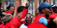 Demonstranten in roten T-shirts recken die Fäuste, in der Mitte ein Mann mit fancy Sonnenbrille