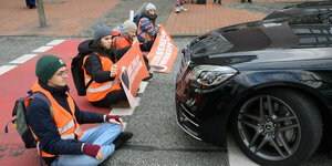 Aktivisten der Letzten Generation sitzen mit Warnwesten aufder Straße, vor ihnen steht ganz nah ein Auto