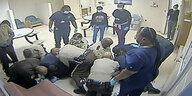 Unscharfes Bild einer Überwachungskamera: Mehrere Uniformierte und Krankenhausmitarbeiter werfen sich auf einen am Boden liegenden Körper in einem Aufenthaltsraum, andere stehen daneben