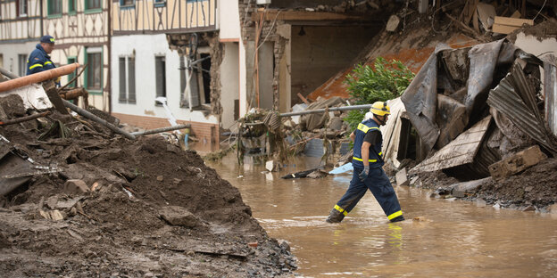 Ein Mann mit Helm läuft durch eine Überflutete Straße. Im Hintergrund zerstörte Häuser