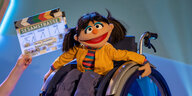 Eine Puppe aus der Sesamstraße mit oranger Jacke und dunklen Haaren sitzt im Rollstuhl