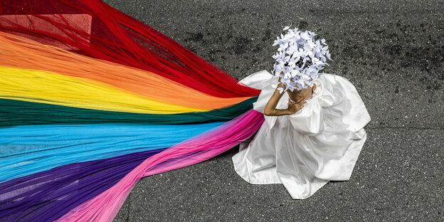 Eine Person mit weißem Rock und weißen Blumen auf dem Kopf trägt einen Regenbogenschleier