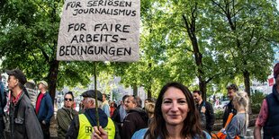 "Für seriösen Journalismus. Für faire Arbeitsbedingungen" stehtauf einem Schild, dass eine Frau mit langen glatten, dunklen Haaren in die Luft hält. Hinter ihr andere Demonstrierende und Bäume mit grünen Blättern.