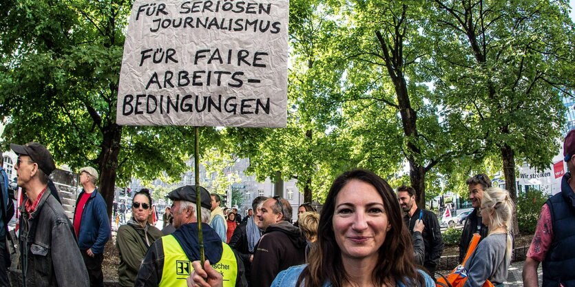 "Für seriösen Journalismus. Für faire Arbeitsbedingungen" stehtauf einem Schild, dass eine Frau mit langen glatten, dunklen Haaren in die Luft hält. Hinter ihr andere Demonstrierende und Bäume mit grünen Blättern.